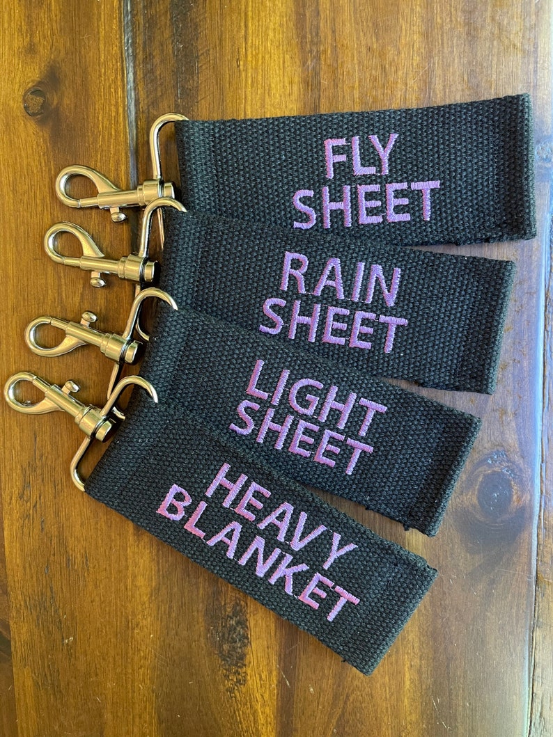 May include: Four black fabric keychains with silver key rings. The keychains are labeled "Fly Sheet", "Rain Sheet", "Light Sheet", and "Heavy Blanket" in purple embroidery.