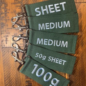 May include: Five green fabric keychains with silver key rings. The keychains are labeled with the text "SHEET", "MEDIUM", "MEDIUM", "50g SHEET", and "100g".