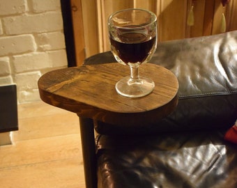 Puede incluir: Una mesa auxiliar con tablero y base de madera marrón oscuro conectados por un tubo de metal negro vertical. La mesa está situada junto a un sofá de cuero negro y sostiene un vaso transparente lleno de un líquido rojo oscuro.