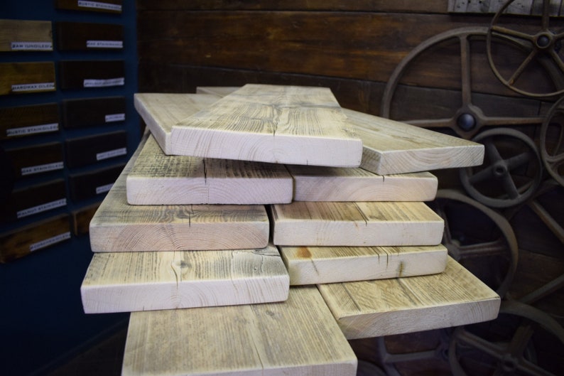 Puede incluir: Una pila de tablones de madera rectangulares de color claro con patrones de veta visibles y textura natural. Las tablas est&aacute;n dispuestas en una pila escalonada, mostrando los bordes crudos y los tonos beige y gris de la superficie.