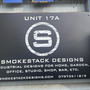 Puede incluir: Un letrero rectangular negro con el texto blanco UNIT 17A, un logotipo circular con una letra S estilizada y las palabras SMOKESTACK DESIGNS. Debajo, se lee INDUSTRIAL DESIGNS FOR HOME, GARDEN, OFFICE, STUDIO, SHOP, BAR, ETC. con sitio web y tel&eacute;fono.