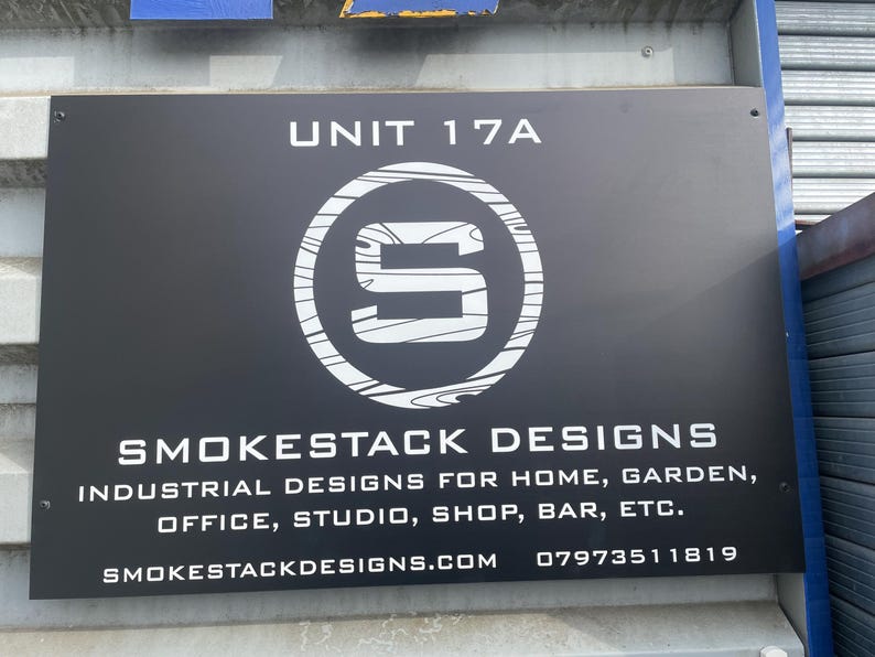 Puede incluir: Un letrero rectangular negro con el texto blanco UNIT 17A, un logotipo circular con una letra S estilizada y las palabras SMOKESTACK DESIGNS. Debajo, se lee INDUSTRIAL DESIGNS FOR HOME, GARDEN, OFFICE, STUDIO, SHOP, BAR, ETC. con sitio web y tel&eacute;fono.