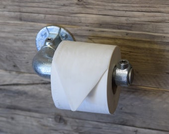 Puede incluir: Un portarrollos de papel higiénico fabricado con tuberías y accesorios de acero galvanizado de estilo industrial. El soporte cuenta con una brida circular montada en la pared y un brazo horizontal que sostiene un rollo de papel higiénico blanco.