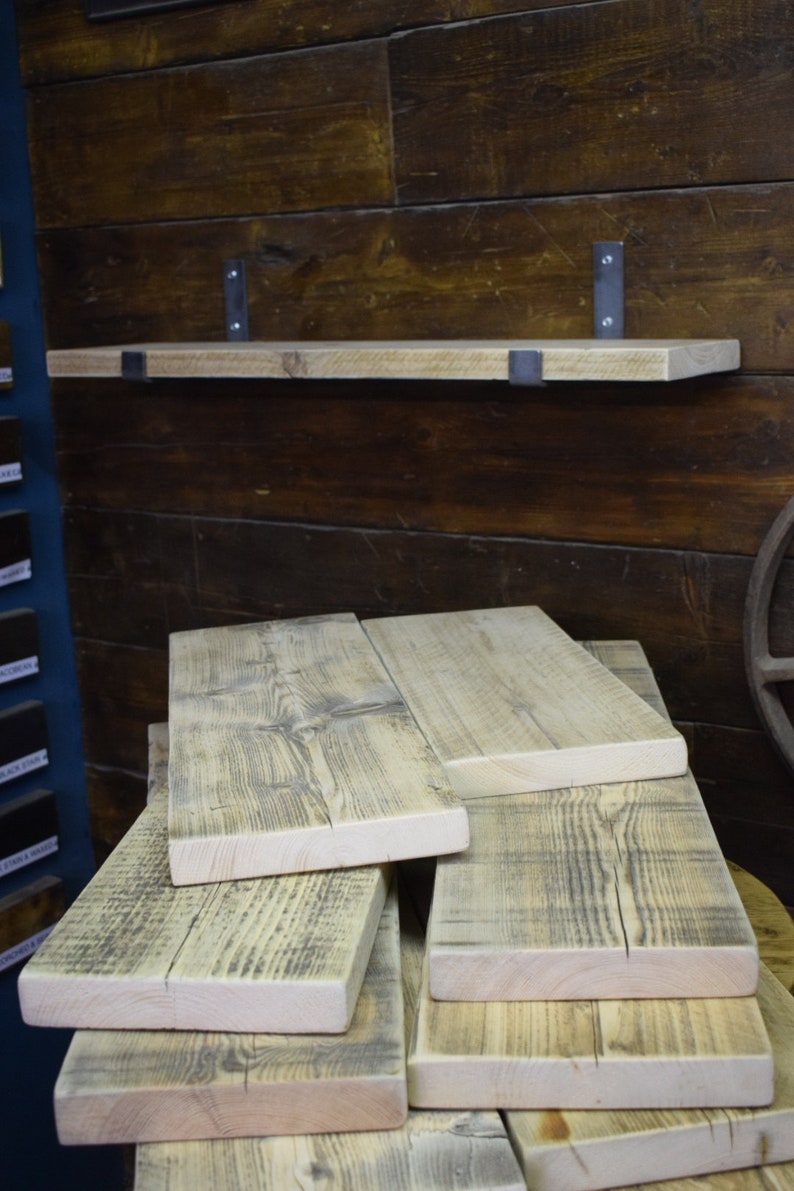 Puede incluir: Un estante de madera clara montado con dos soportes met&aacute;licos en una pared de madera oscura, con varias tablas rectangulares adicionales apiladas en primer plano, cada una con un patr&oacute;n de veta visible y una textura natural sin tratar.