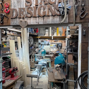 Puede incluir: Un taller de carpinter&iacute;a con un letrero de metal que dice SMOKESTACK DESIGNS, varios ganchos de metal, una cuerda y un adorno en forma de coraz&oacute;n. El espacio contiene herramientas el&eacute;ctricas, sierras ingletadoras, madera y un extintor rojo.