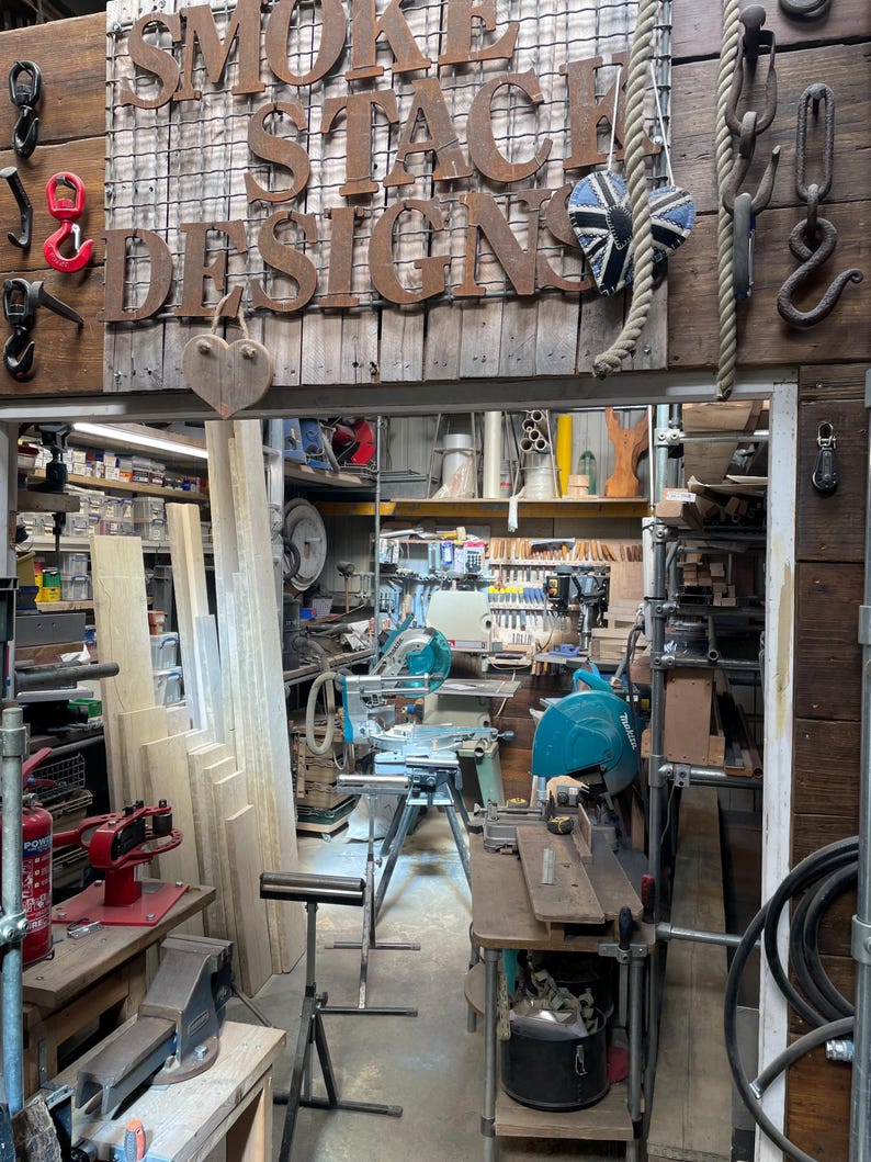 Puede incluir: Un taller de carpinter&iacute;a con un letrero de metal que dice SMOKESTACK DESIGNS, varios ganchos de metal, una cuerda y un adorno en forma de coraz&oacute;n. El espacio contiene herramientas el&eacute;ctricas, sierras ingletadoras, madera y un extintor rojo.