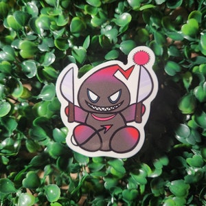 May include: A black and red cartoon character with a menacing grin, holding two knives. The character has a red star on its head and is sitting on a white background.