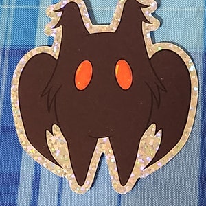 Może przedstawiać: Czarno-czerwona naklejka z brokatem przedstawiająca kreskówkową ćmę z dużymi, okrągłymi oczami. Ćma ma zaokrąglone ciało i duże skrzydła.