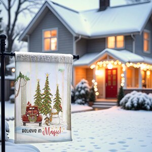 May include: A winter garden flag with a red truck, Christmas trees, a snowman, and the text "Believe in Magic!". The flag is set against a snowy backdrop with a house in the background. The flag is approximately 46 cm wide and 30 cm tall.