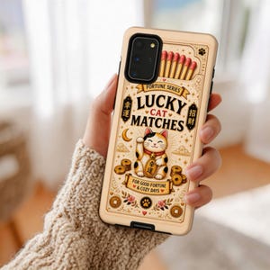 May include: A beige phone case featuring a Lucky Cat design with the text "Lucky Cat Matches." The case has a matchbox design with red match heads, gold coins, and floral accents. The phone case is held in a person's hand.