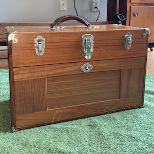 May include: A vintage wooden toolbox with a brown leather handle, metal clasps, and corner protectors. The toolbox is set on a green rug, with a drafting table and wooden cabinet in the background. The toolbox is a warm brown color.