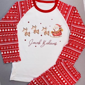 May include: A two-piece pajama set featuring a white top with red sleeves and matching red pants. The top has a Santa and reindeer graphic with the text "Jacob Believes". The sleeves and pants have a red and white pattern with reindeer and snowflake designs.