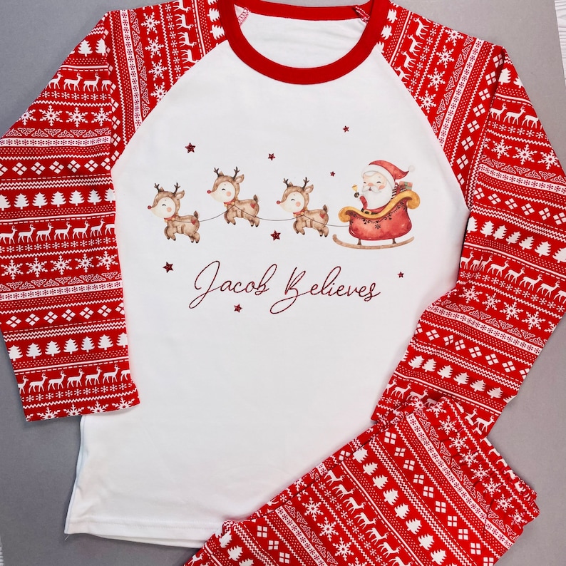 May include: A two-piece pajama set featuring a white top with red sleeves and matching red pants. The top has a Santa and reindeer graphic with the text "Jacob Believes". The sleeves and pants have a red and white pattern with reindeer and snowflake designs.
