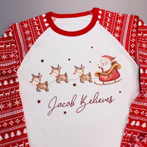 May include: White and red Christmas pajamas with a festive pattern on the sleeves and pants. The shirt features a Santa Claus in a sleigh pulled by reindeer, with the text "Jacob Believes".