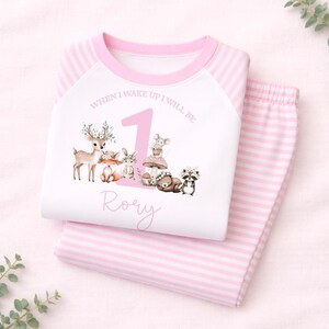 May include: A white and pink striped pajama set featuring a graphic of a large number 1, woodland animals, and the text "WHEN I WAKE UP I WILL BE" and the name "Rory" printed on the front of the long-sleeved shirt with raglan sleeves.