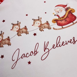 May include: A festive illustration featuring Santa Claus in a red sleigh pulled by reindeer. The image includes the text "Jacob Believes" in a glittery red script, along with red star accents, all set against a white background. A charming Christmas design.