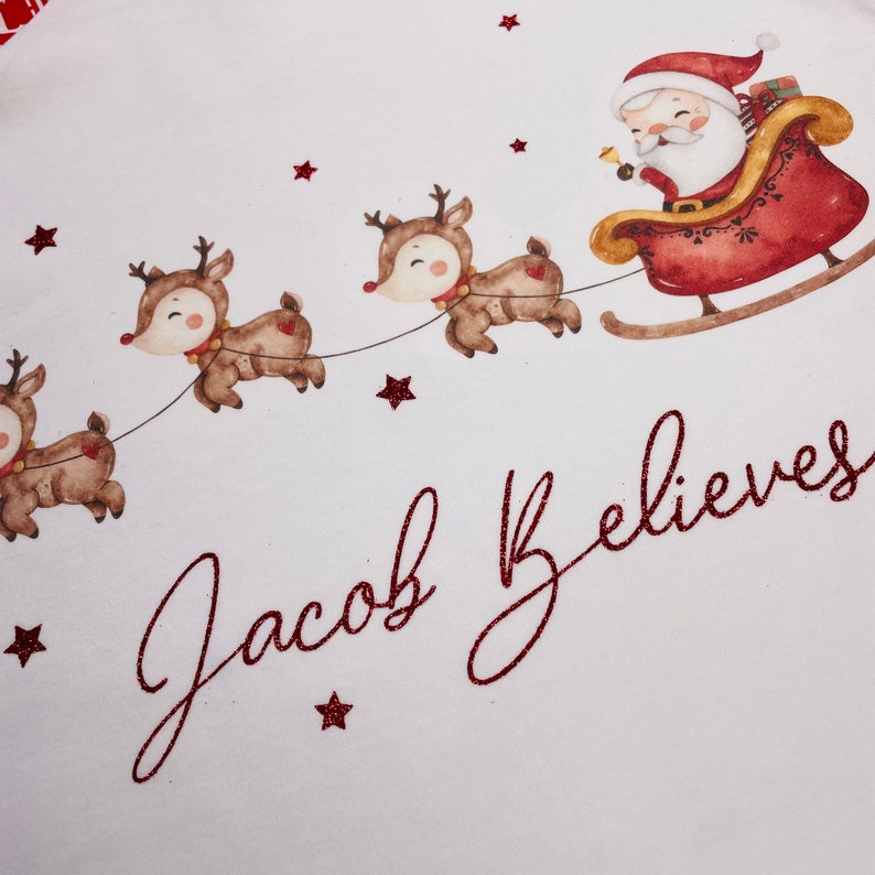 May include: A festive illustration featuring Santa Claus in a red sleigh pulled by reindeer. The image includes the text "Jacob Believes" in a glittery red script, along with red star accents, all set against a white background. A charming Christmas design.