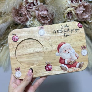 May include: A rectangular wooden serving board featuring a circular cutout, decorated with illustrations of Christmas ornaments, snowflakes, and a Santa Claus holding a mug and cookies, with the text Santa, A little treat for you, Love.