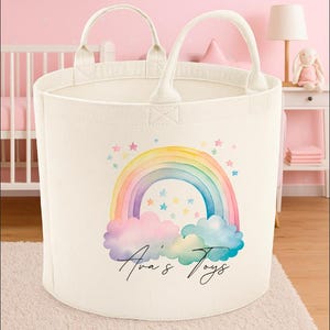 May include: Off-white canvas toy basket with two handles. The front features a watercolor rainbow design with pink, yellow, blue, and purple hues, along with the text "Ava's Toys". The basket is cylindrical and sits on a light pink rug.