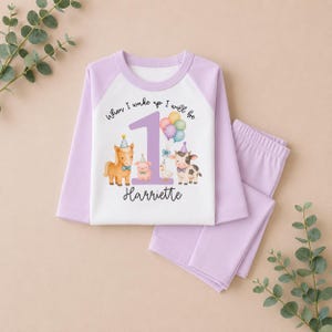 May include: A long-sleeve pajama set with light purple sleeves and pants. The white shirt features a large purple number 1, a cluster of colorful balloons, and illustrations of a horse, pig, duck, and cow. Text reads When I wake up I will be 1 Harriette.