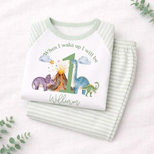 May include: A white long-sleeve shirt with light green striped sleeves and a matching pair of striped pants. The shirt features a graphic of a volcano, dinosaurs, and the text When I wake up I will be 1 William in green lettering.