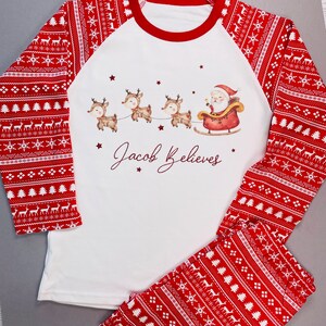 May include: A white and red Christmas pajama set. The top has a red neckline and sleeves with a festive pattern. The front features Santa in a sleigh pulled by reindeer, with the text "Jacob Believes". The pants have a matching pattern.
