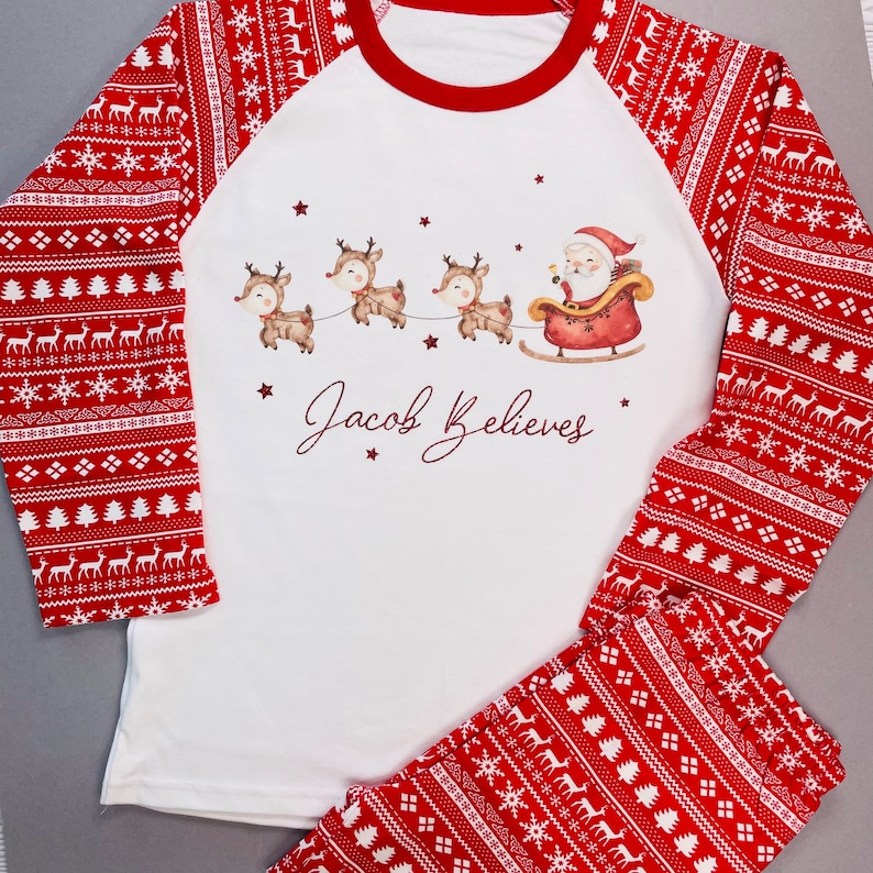 May include: A white and red Christmas pajama set. The top has a red neckline and sleeves with a festive pattern. The front features Santa in a sleigh pulled by reindeer, with the text "Jacob Believes". The pants have a matching pattern.