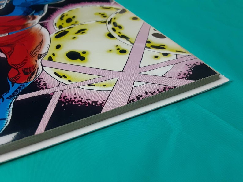 May include: Close-up of the spine of a comic book with a colorful illustration of a superhero in a red, blue, and white costume standing in front of a pink and yellow background.