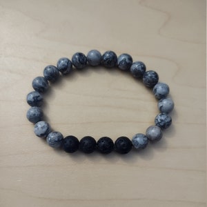 May include: A gray and black beaded bracelet with round beads. The bracelet is made with gray marbled beads and black lava rock beads.