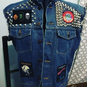 May include: A blue denim vest with silver studs and various patches. The patches include a pumpkin, a skull, a green monster, a band logo, and a movie title. The vest has a pocket on the front.