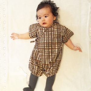 May include: A baby wearing a brown and white plaid romper with a brown sash. The romper has a loose fit and is made of a soft, comfortable fabric.
