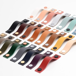 May include: A collection of 20 leather cabinet pulls in various colours, arranged in a staggered pattern. Each pull has a curved shape and two metal screws.