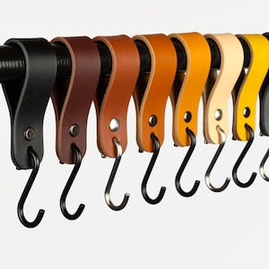 May include: A set of ten leather hooks in various colours, including black, brown, tan, yellow, and white. The hooks are attached to a black metal rod and have a simple, minimalist design.