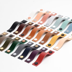 Könnte beinhalten: Eine Auswahl an Leder-Schrankgriffen in verschiedenen Farben, die in Fächerform angeordnet sind. Die Griffe sind aus Leder gefertigt und haben eine geschwungene Form. Die Griffe sind alle in verschiedenen Farben, darunter Braun, Grün, Blau, Rot und Gelb.