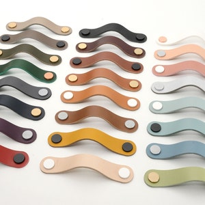 May include: A set of 16 leather cabinet pulls in various colors, including brown, red, green, blue, and gray. Each pull has a curved shape and is accented with a metal button.