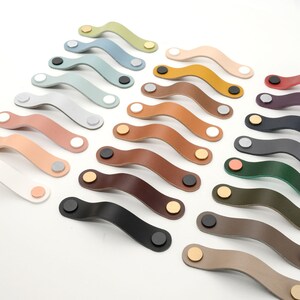 May include: Assortment of 18 leather cabinet pulls in various colors, including green, brown, black, white, and beige. Each pull has a curved shape and features a gold metal screw.