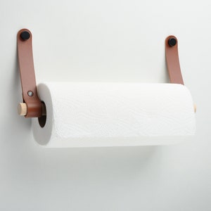 May include: A brown leather paper towel holder with wooden dowels and black metal hardware mounted on a white wall.