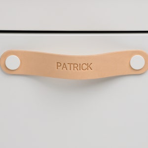 Peut inclure: Une poignée de tiroir en cuir brun clair avec des boutons ronds blancs à chaque extrémité. La poignée porte le nom "PATRICK" en relief sur le cuir.