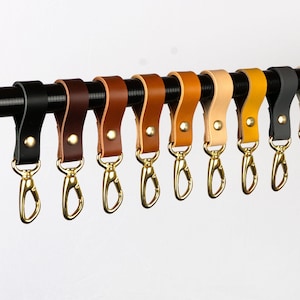 Könnte beinhalten: Eine Sammlung von Leder-Schlüsselanhängern in verschiedenen Farben, darunter Schwarz, Braun, Beige und Gelb. Jeder Schlüsselanhänger hat einen goldfarbenen Verschluss und ist an einer schwarzen Stange befestigt. Die Schlüsselanhänger werden vor weißem Hintergrund präsentiert.