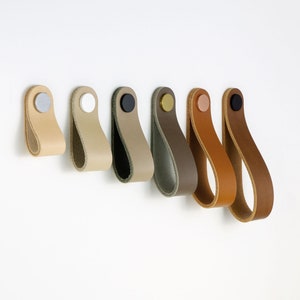 Może przedstawiać: Zestaw siedmiu skórzanych uchwytów do szafek w różnych kolorach, w tym czarny, brązowy, beżowy i szary. Każdy uchwyt ma metalowy gałkę w innym kolorze.