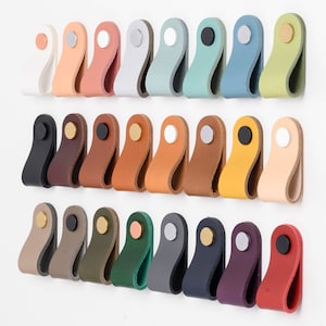 Op de afbeelding: Assortiment van 24 leren kastgrepen in verschillende kleuren, waaronder zwart, bruin, wit, groen, blauw, rood en beige. Elke greep heeft een ronde metalen knop.