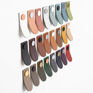 Op de afbeelding: Een verzameling van 24 lederen ladegrepen in verschillende kleuren, waaronder wit, zwart, bruin, rood, groen, blauw en geel. Elke greep heeft een ronde metalen knop in het midden.