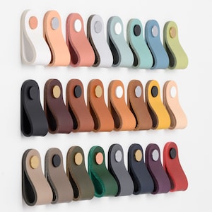 Op de afbeelding: Een verzameling van 20 lederen ladegrepen in verschillende kleuren, waaronder zwart, bruin, beige, groen, blauw, rood en wit. Elke greep heeft een eenvoudig, gebogen ontwerp en een metalen schroef.