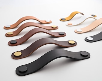 May include: Assortment of leather cabinet pulls in various colors, including brown, black, white, and beige. Each pull features a curved design with a metal accent.