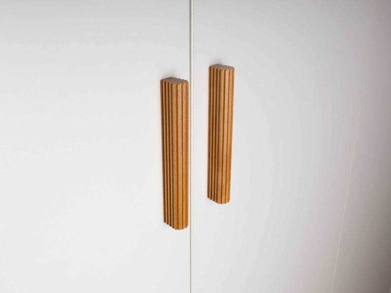 Op de afbeelding: Twee houten kastgrepen met een verticaal geribbeld ontwerp. De grepen zijn lichtbruin van kleur en zijn gemonteerd op een witte kastdeur. De grepen zijn rechthoekig met een afgeronde bovenkant.