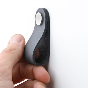 Op de afbeelding: Een zwarte leren kastgreep met een zilveren metalen schroef. De greep is lusvormig en is ontworpen om als handvat te worden gebruikt.