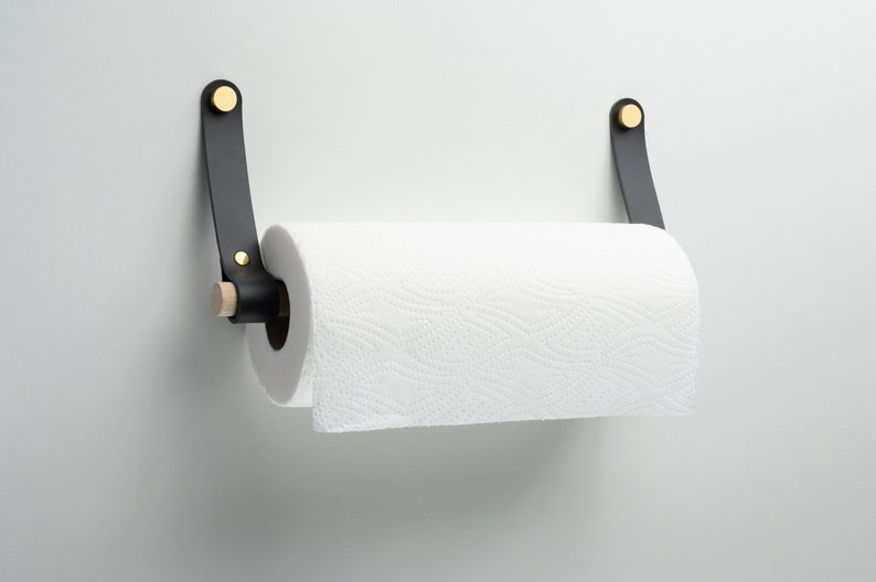 May include: Black leather paper towel holder with brass accents and a wooden dowel. The holder is mounted on a white wall.