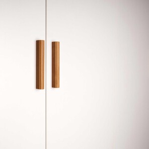 Op de afbeelding: Twee verticale, geribbelde houten kastgrepen zijn gemonteerd op een witte kastdeur. De grepen zijn warm lichtbruin van kleur en hebben een cilindrische vorm. De kastdeur is gesloten.