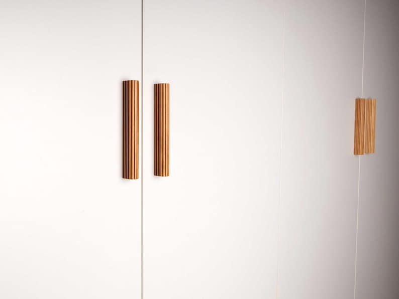 Op de afbeelding: Twee verticale, geribbelde houten kastgrepen zijn gemonteerd op een witte kastdeur. De grepen zijn warm lichtbruin van kleur en hebben een cilindrische vorm. De kastdeur is gesloten.