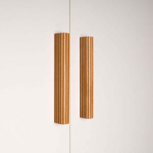 Op de afbeelding: Twee verticale, lichtbruine houten kastgrepen met een geribbeld ontwerp zijn gemonteerd op een witte kastdeur. De grepen zijn rechthoekig en hebben een warme, natuurlijke houttint. De kastdeur is gesloten.